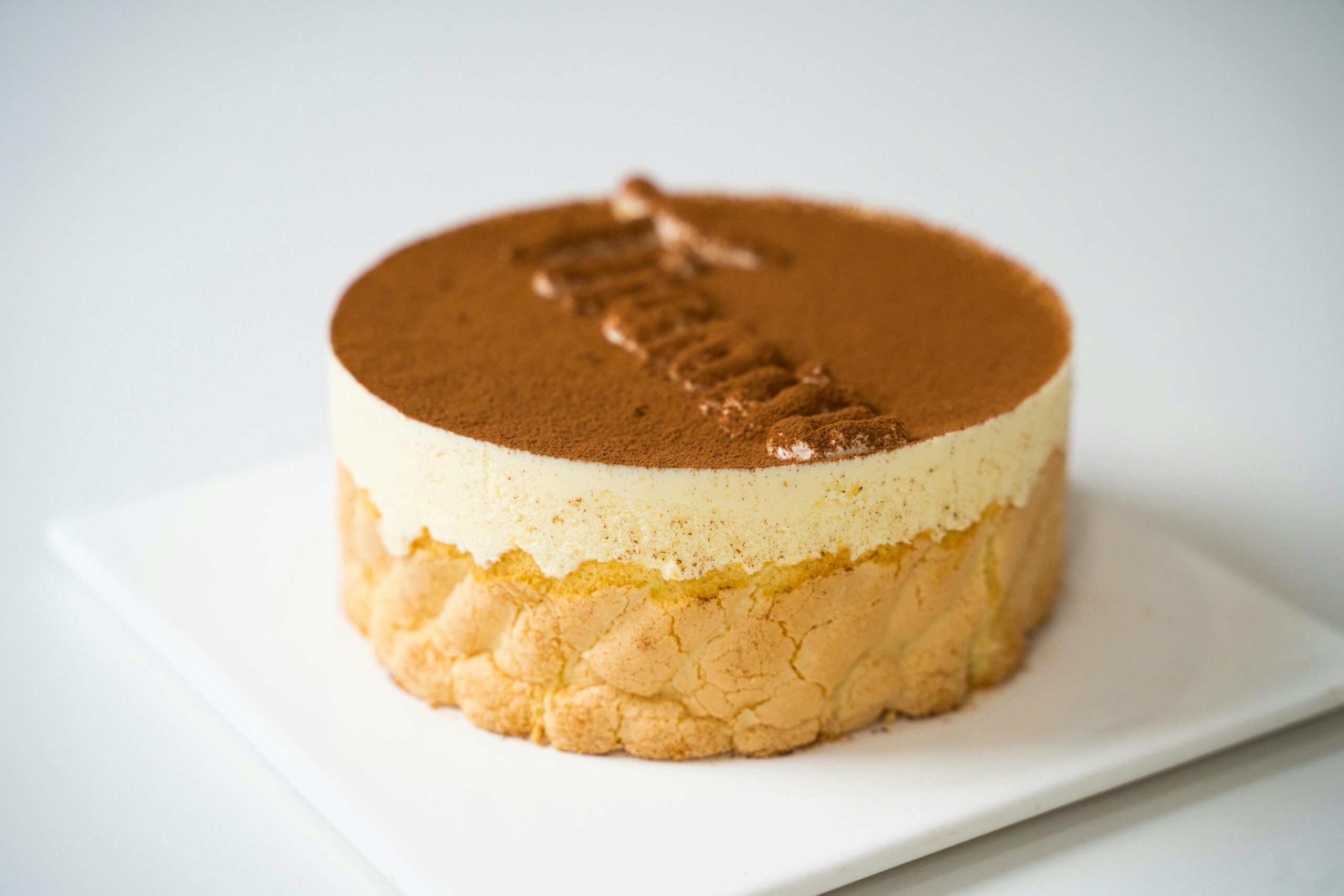 Tiramisu cake placed on a white plate. It's written tiramisu on top of the cake with cocoa dusting on top.
