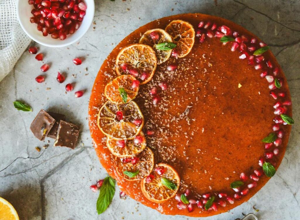 this image shows a brown orange cake with orange slices, some leaves and pomegranate seeds on top of it.