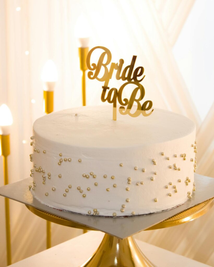 vannilla cake for briday shower. A bride to be stick on the cake, sides of cake decorated with golden sugar beads.