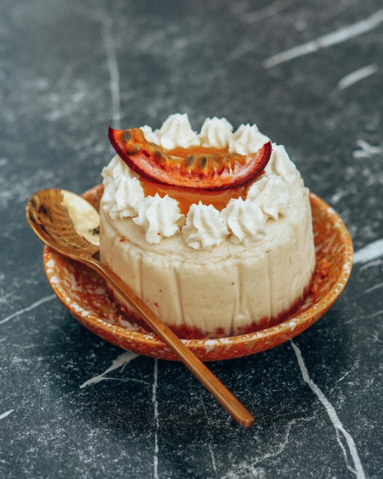 small passion fruit cake, topped with passion fruit pure. decorated with a piece of passion fruit on top. its placed on a plate with a spoon.
