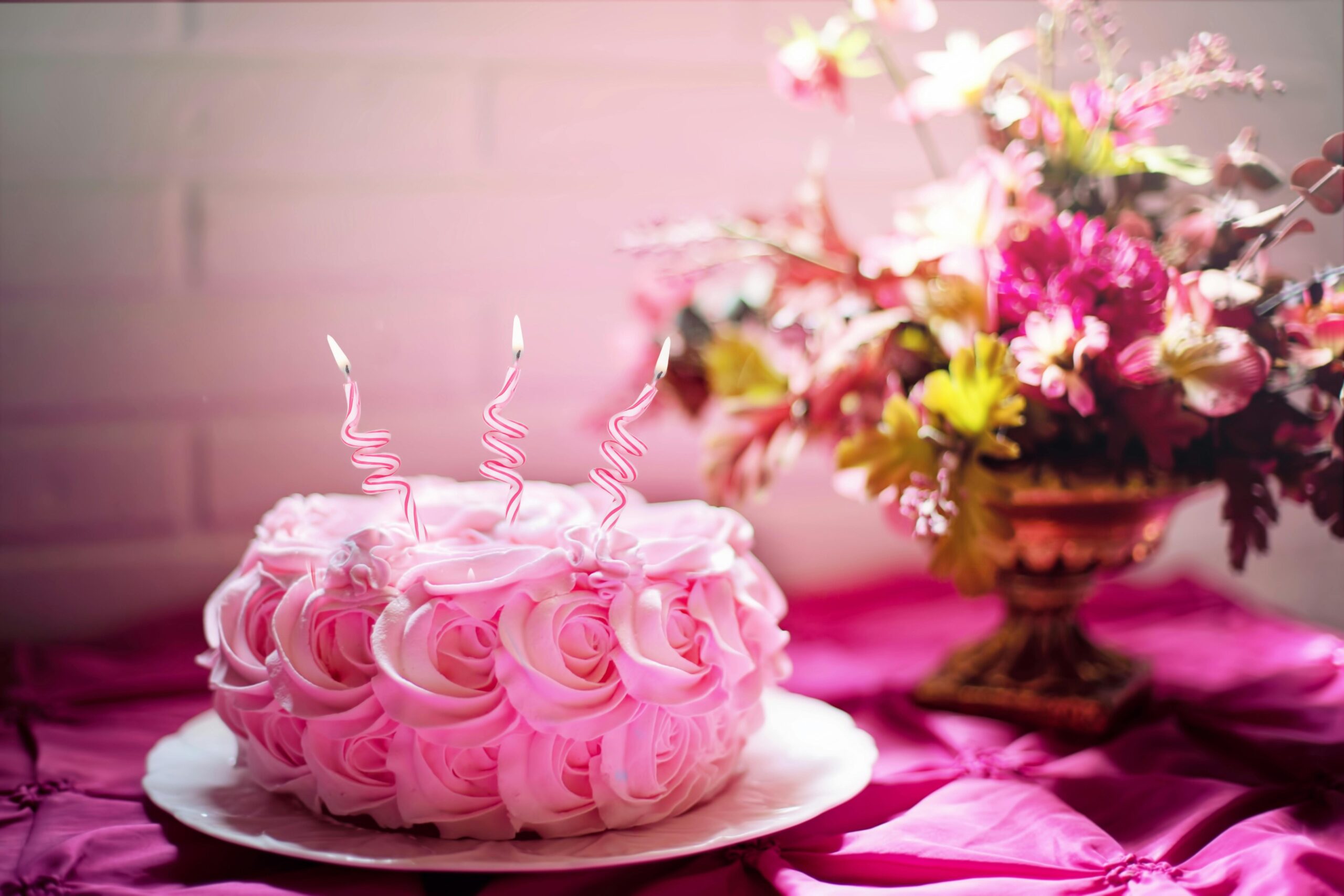 a celebration cake placed on a whie plate. The cake is covered in pink rose decorations.. Three pink spiral candles are placed on the top.