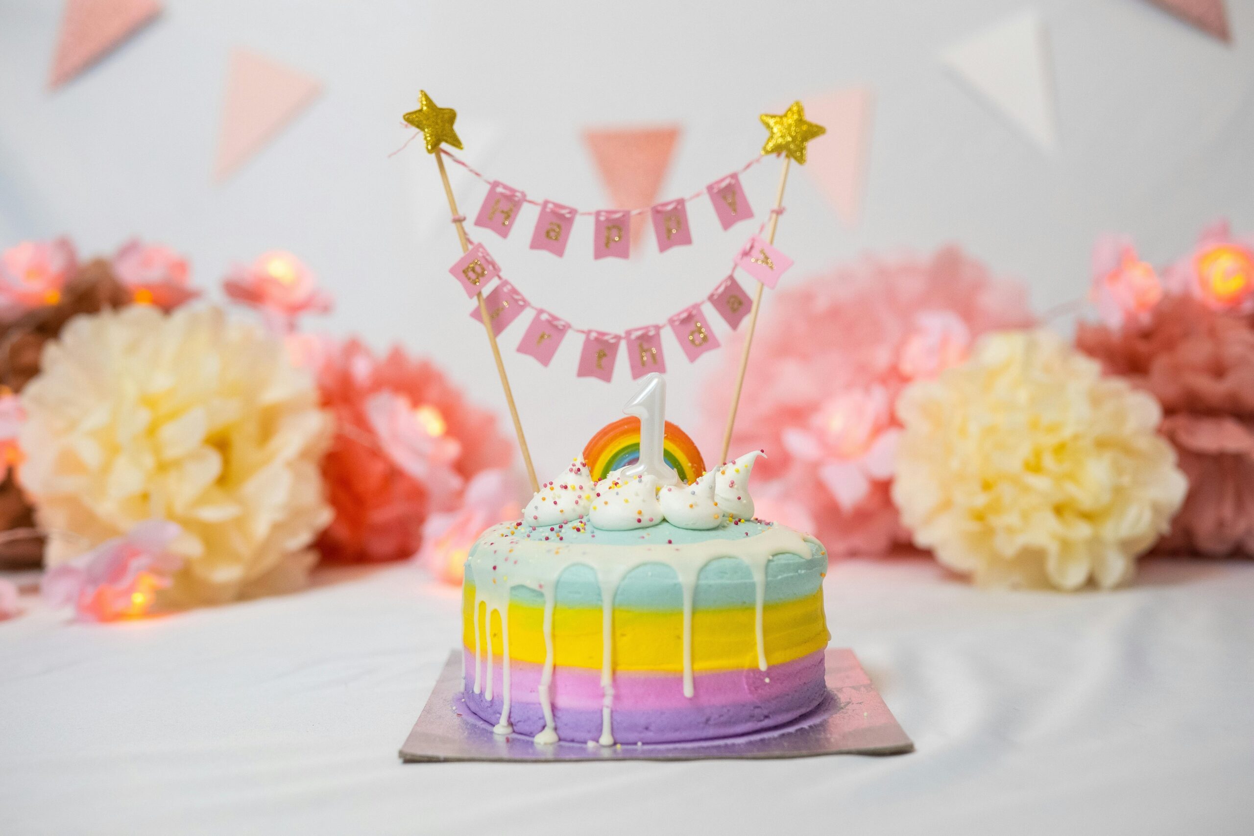 Bakery - a rainbow themed three tiered birthdaycake for one year old. the three layers of cakes are colored in violet, yellow and green. A happy birthday lettered banner placed between two sticks with a star on the top is placed on the top of the cake along with no.1 candle and a small rainbow.