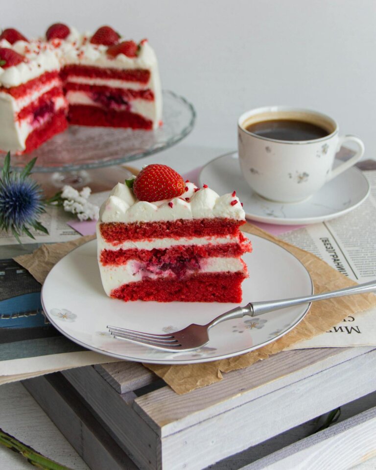 Red velvet cake topped with strawberries and a piece of the cake kept on a plate that shows the layers, a mug of coffee kept along with the cake.