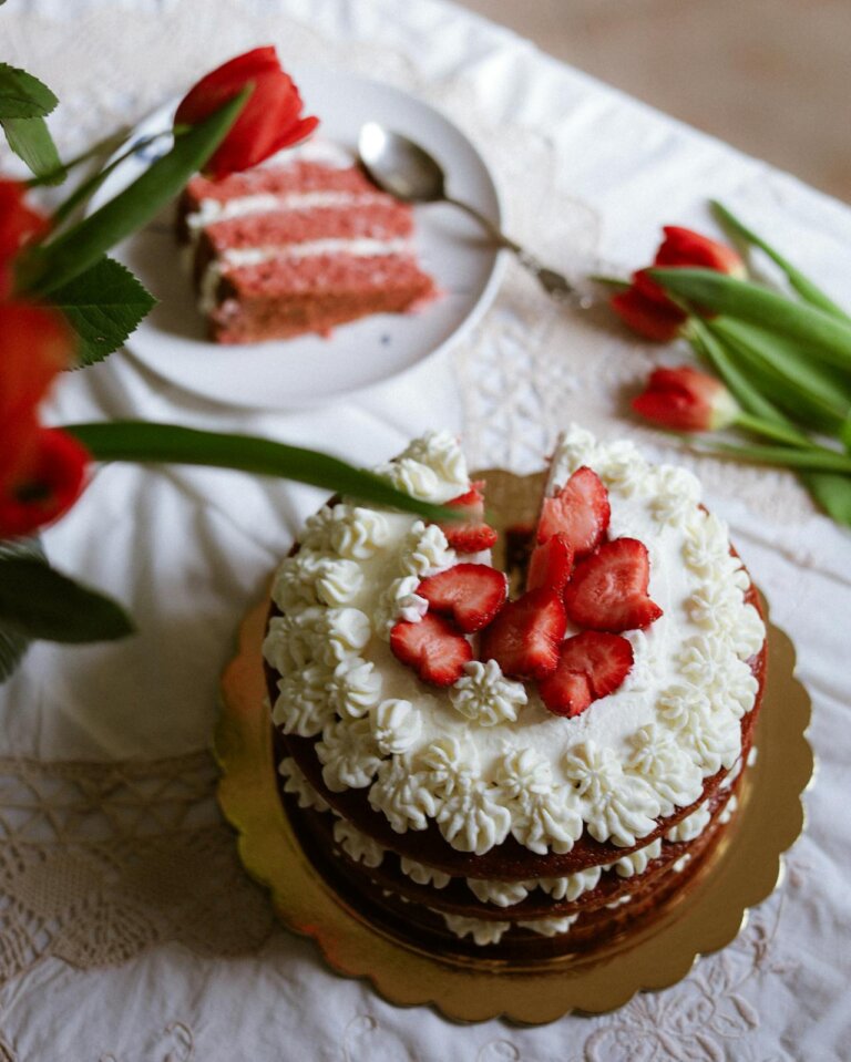 Vanilla strawberry cake made and topped with fresh strawberries. It has whipped cream frosting. one piece is kept aside on a plate with a spoon, some red tulips on the table.