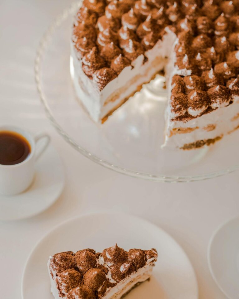 A tiramisu cake placed on a table with choco powder on top, a cup of coffee near the cake. A piece is cut and kept aside on a plate.