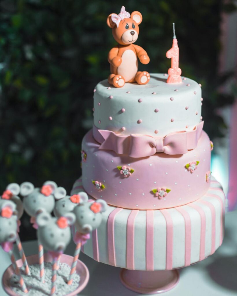 Bakery -A blue and pink themed three tiered birthday cake for a one year old, an edible teddy bear placed on top of the cake, a pink bow tie tied between the second and third tier.